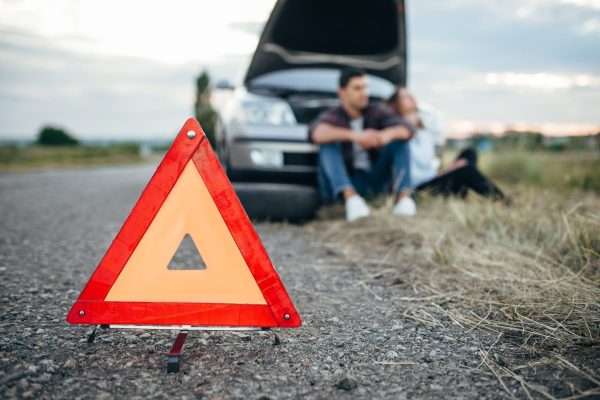 Broken car concept, man sitting on tire against breakdown triangle on asphalt road. Problem with vehicle, warning sign