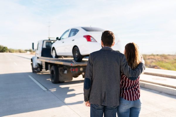 Rear view of a couple hugging and seeing a tow truck go down the road with their broken-down car